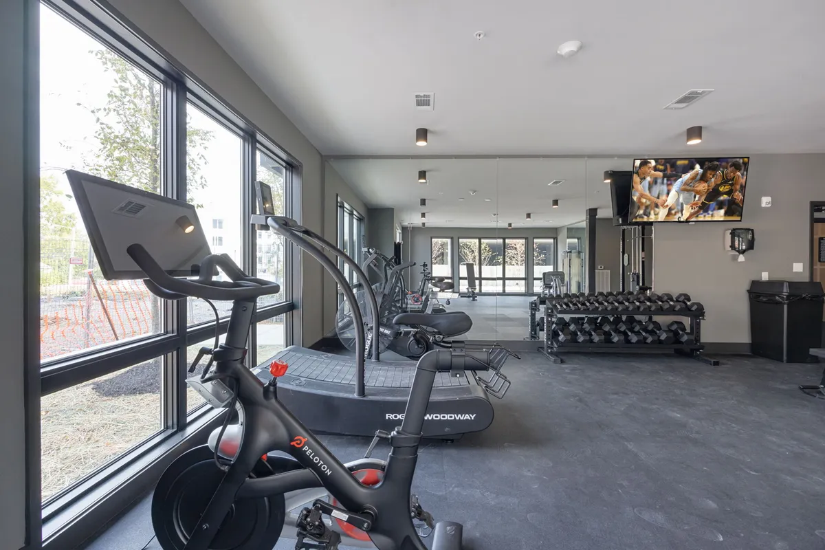 A modern fitness center featuring a Peloton bike, treadmill, dumbbell rack, and large windows for natural light. The space is clean, well-equipped, and designed for both personal and group workouts.