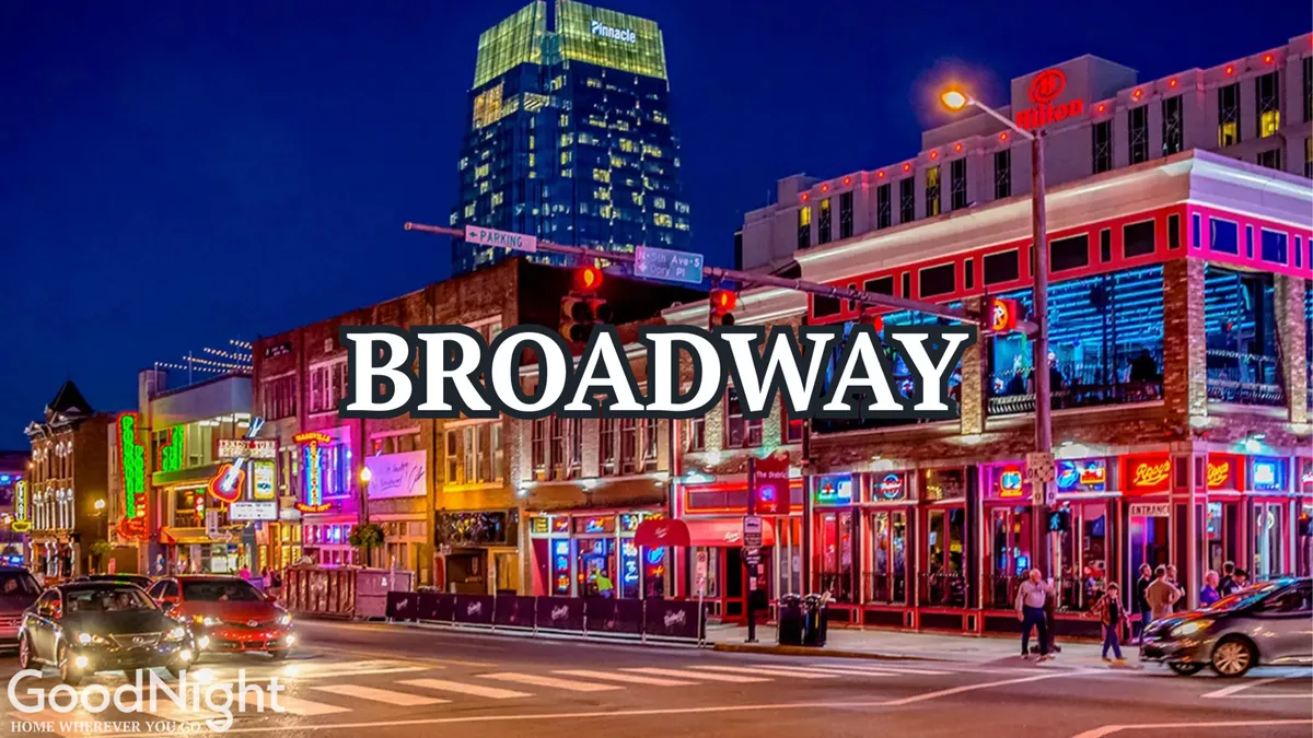 A lively stretch of Nashville's Broadway at night, brimming with neon signs, bustling crowds, and vibrant city energy. People stroll the sidewalks, taking in music, nightlife, and the iconic character of this entertainment district.
