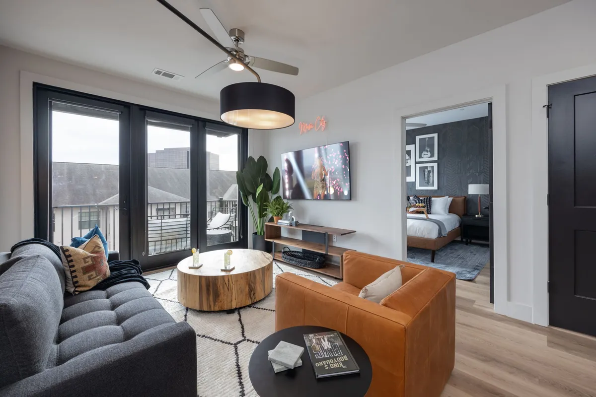 A stylish, sunlit living room with a plush sofa, modern coffee table, large television, and vibrant green plant. The space exudes warmth and comfort, perfect for relaxing, entertaining, or enjoying a cozy movie night. Contemporary decor and floor-to-ceiling windows create an inviting, airy atmosphere.