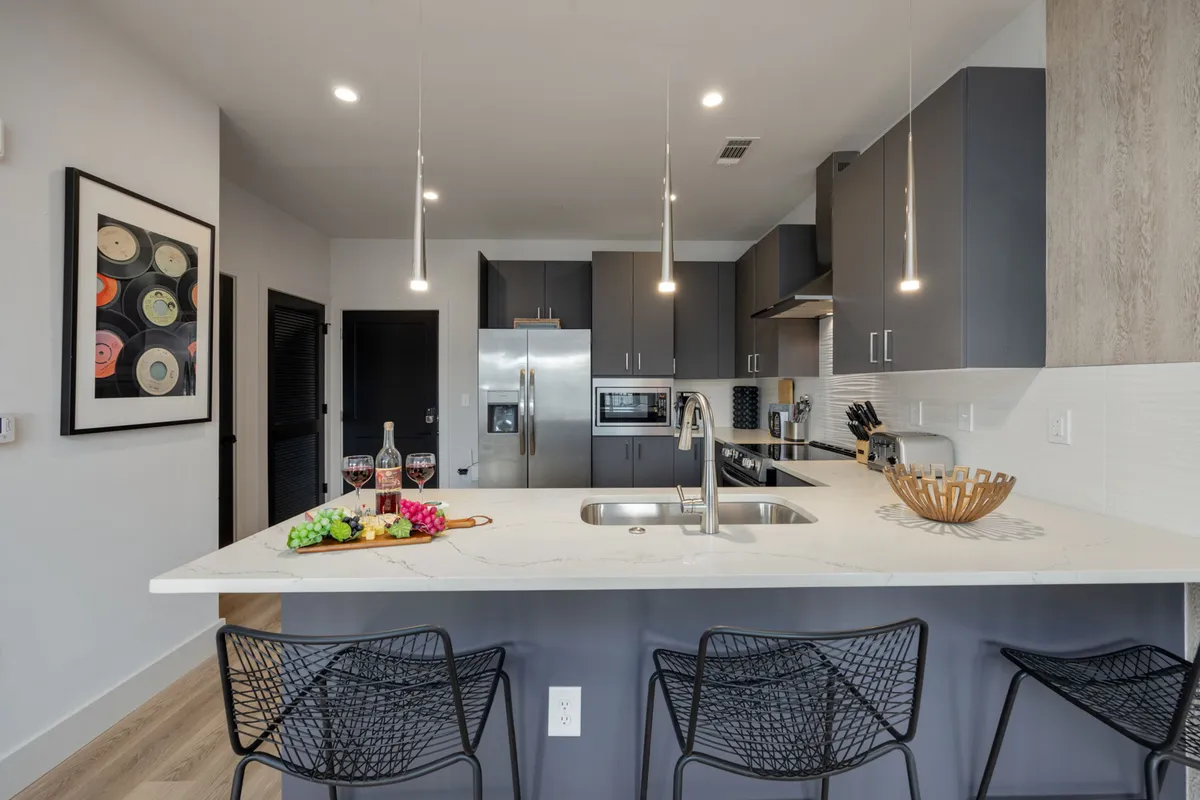 A warm and inviting kitchen scene with a center island lined with bar stools, a wine and fruit arrangement, modern kitchen appliances, and colorful artwork on the wall. The area looks ideal for entertaining and casual meals.