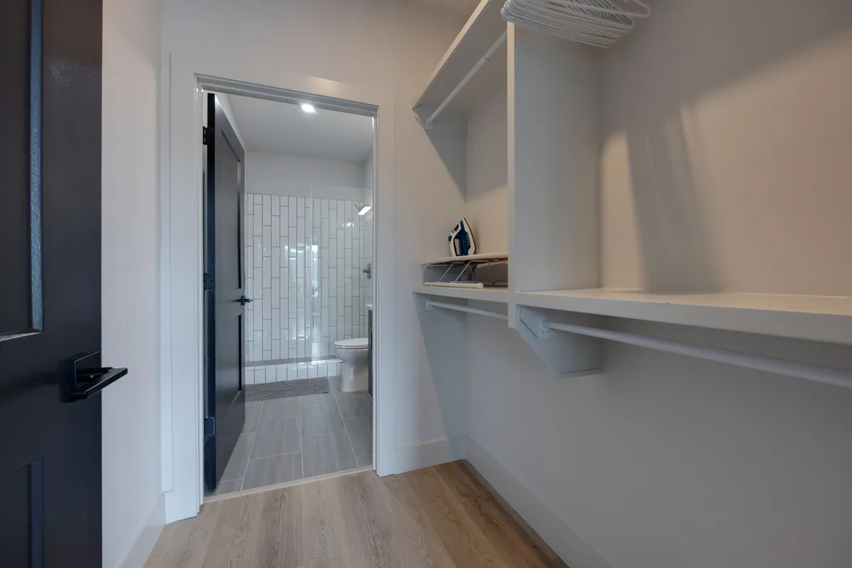 A functional area with an open closet, visible doorway to a bathroom, and an ironing board. The setup presents an organized space perfect for storage and easy morning routines, echoing the walk-in closet and ensuite bath features.
