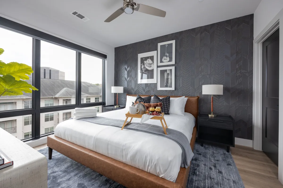 A sunlit bedroom featuring a plush bed topped with decorative pillows, musical-themed wall art, large windows streaming in natural light, and stylish bedside lamps. A tray with breakfast items sits on the bed, suggesting a leisurely, comfortable morning.